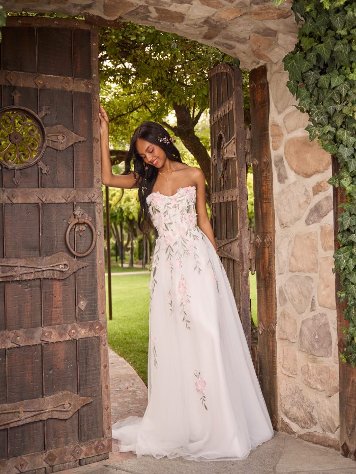 Bride standing in a garden archway wearing a soft A-line wedding dress adorned with pink floral appliqués and embroidered greenery. The gown features a strapless sweetheart neckline and ethereal tulle skirt, creating a whimsical, garden-inspired bridal look.