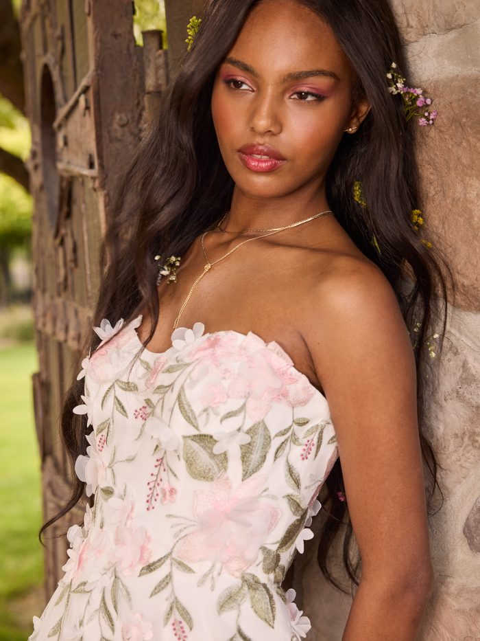 Bride wearing a romantic strapless A-line wedding dress with pink and ivory 3D floral appliqués and soft greenery embroidery. She poses against a rustic stone wall with flowers in her hair, showcasing the gown’s dreamy floral details and sweetheart neckline.