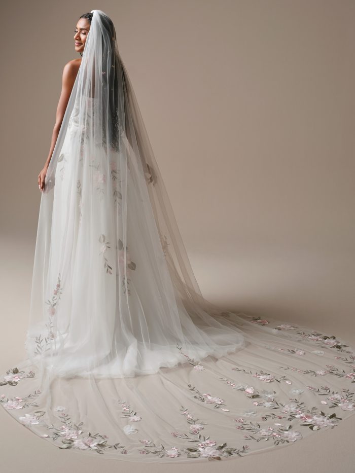 Bride wearing a cathedral-length veil adorned with soft pink and green floral embroidery. The sheer tulle veil flows elegantly over a matching floral lace gown, creating a romantic, garden-inspired bridal look.
