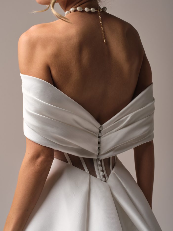 Back view of Mikado A-line bridal gown with off-the-shoulder wrap detail, covered buttons, and sheer illusion corset back, creating a modern yet timeless bridal look.