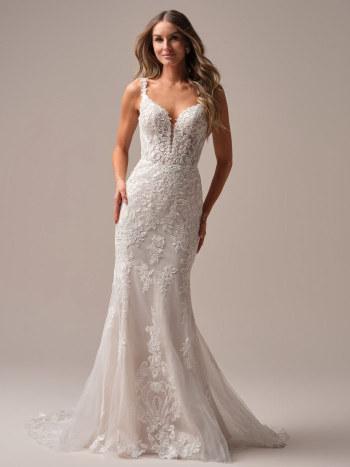 Bride wearing the Rebecca Ingram Candice sheath wedding dress featuring a plunging V-neckline, illusion lace bodice, and floral lace motifs cascading over a fitted silhouette with a soft train.