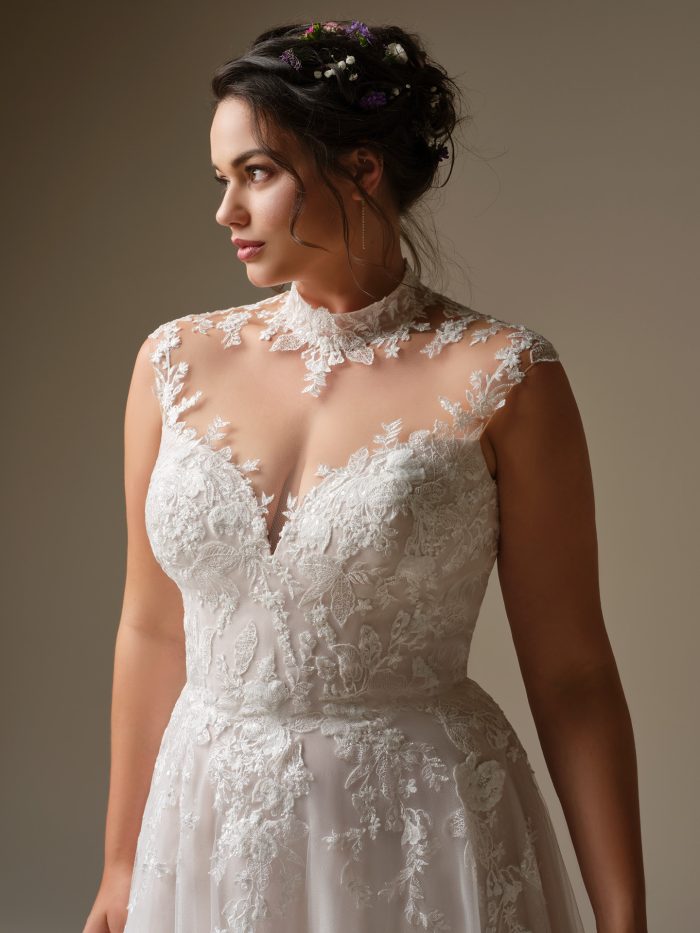 Close-up of the Rebecca Ingram Cody A-line wedding dress highlighting the illusion high neckline, intricate floral lace appliqués, and sheer plunge detail, styled with a floral hairpiece.
