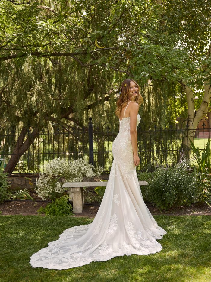 Bride in Rebecca Ingram Eden gown showcasing a low illusion back, cascading floral lace details, and a long chapel train, smiling as she looks over her shoulder in a lush garden.