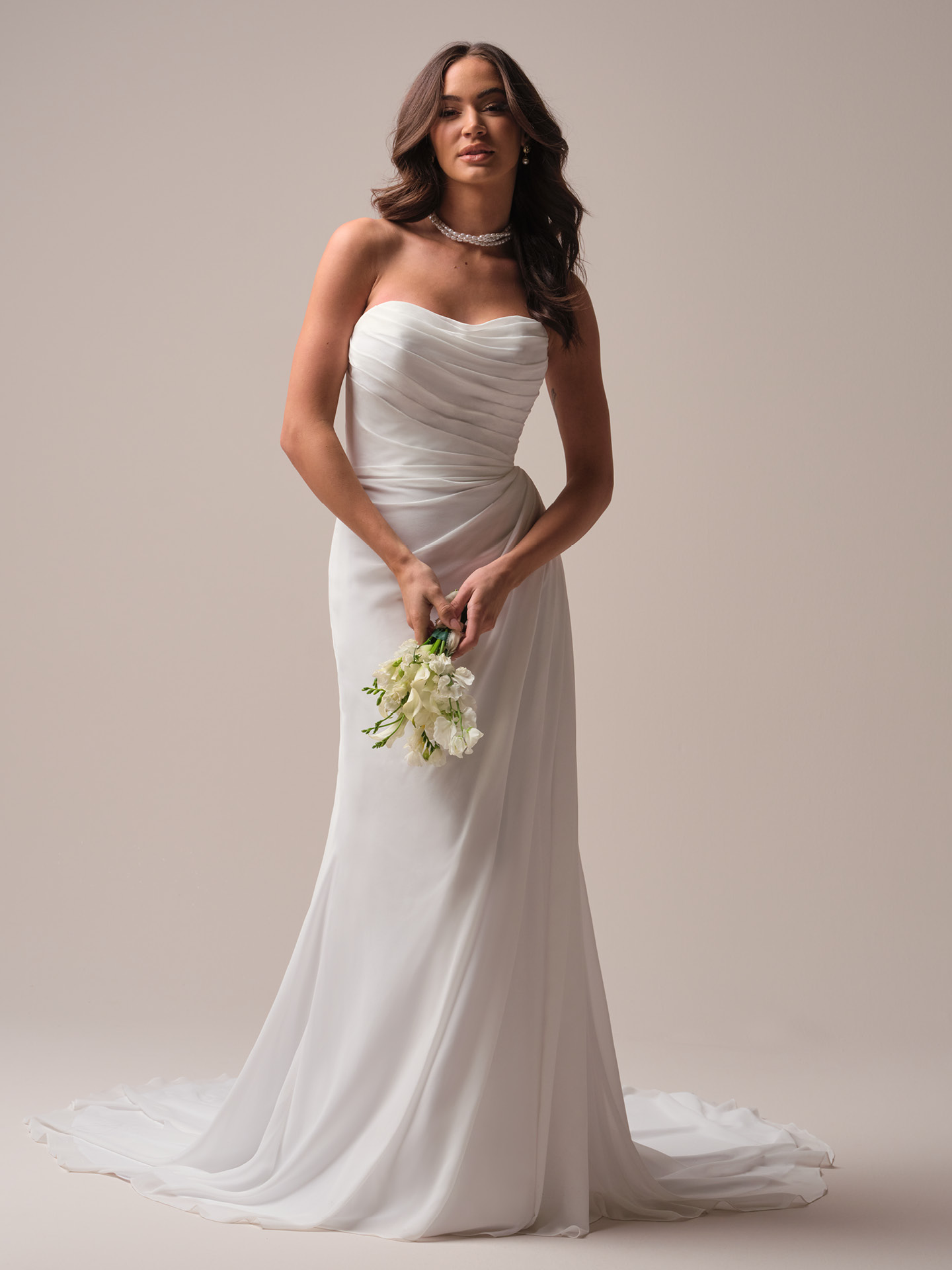 Bride wearing Francesca by Rebecca Ingram, a strapless soft scoop neckline wedding dress with asymmetrically draped bodice and flowing satin skirt, holding a white bouquet against a neutral backdrop.