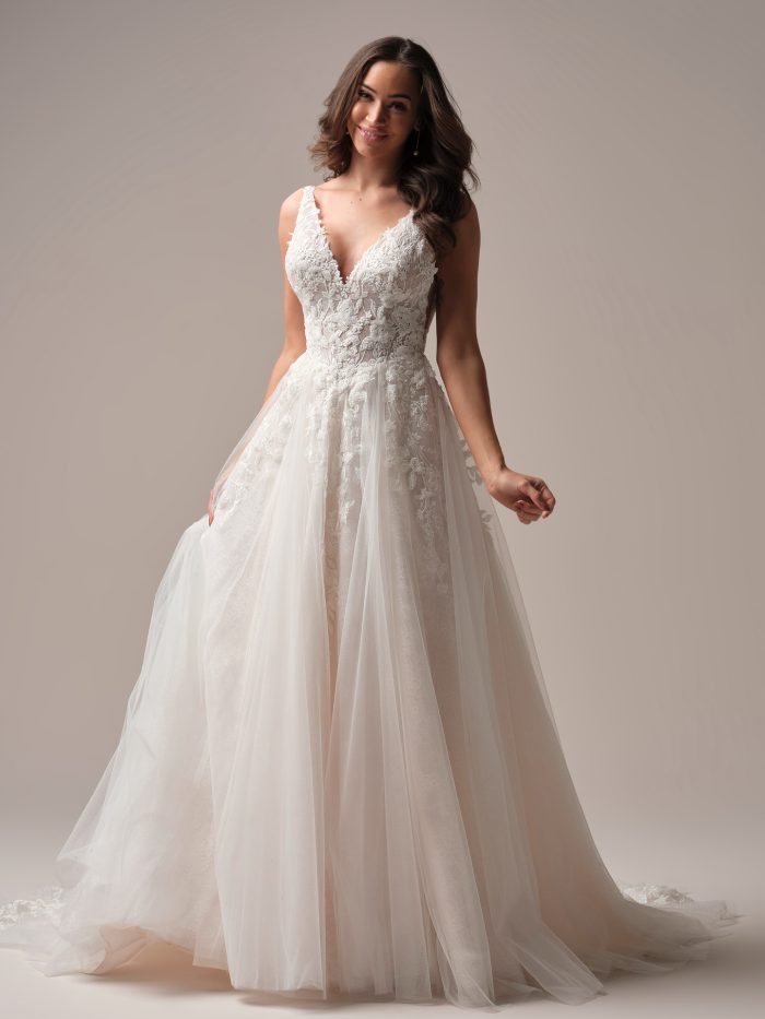 Bride wearing the Rebecca Ingram Honor A-line wedding dress featuring a deep V-neckline, illusion lace bodice with floral appliqués, and a flowing layered tulle skirt for a romantic textured bridal look.
