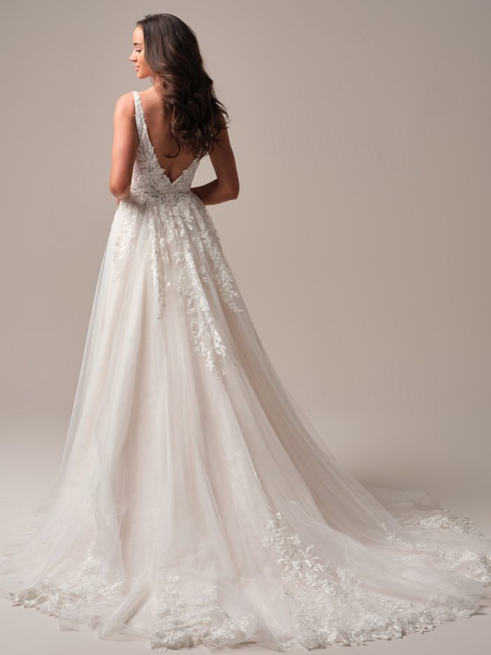 Back view of the Rebecca Ingram Honor A-line wedding dress showcasing a low scoop back, delicate lace straps, and trailing floral appliqués cascading into a chapel-length train.