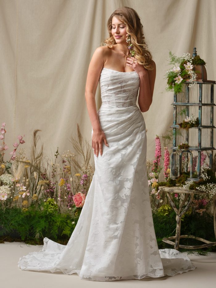 Front view of Rebecca Ingram Kendall wedding dress, a strapless sheath gown featuring ruched tulle over a corset-style bodice and floral jacquard fabric, shown with a romantic garden backdrop.