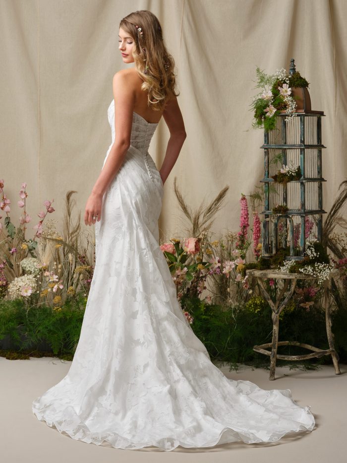 Back view of Rebecca Ingram Kendall wedding dress, showcasing the strapless bodice, button closure, and flowing floral jacquard train against a soft garden-inspired setting.