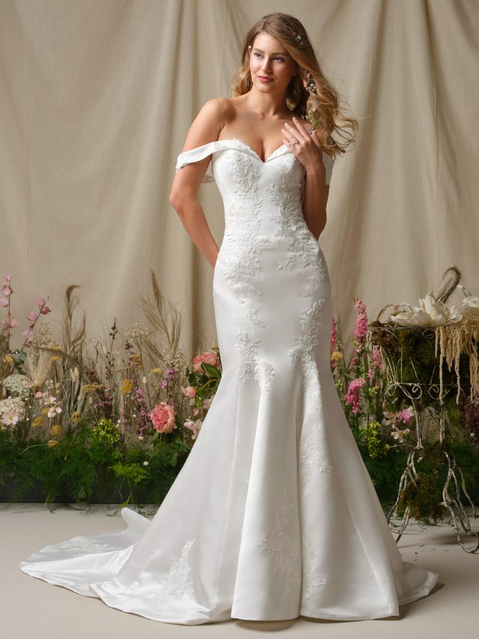 Bride wearing the Rebecca Ingram Miley fit-and-flare wedding dress featuring an off-the-shoulder neckline, floral lace appliqués, and a curve-hugging silhouette with chapel-length train.