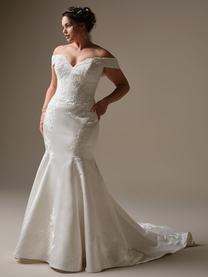 A model wearing the Rebecca Ingram Miley wedding dress, featuring an off-the-shoulder neckline, embroidered lace bodice and a sleek fit-and-flare satin skirt with floral appliqué detailing.