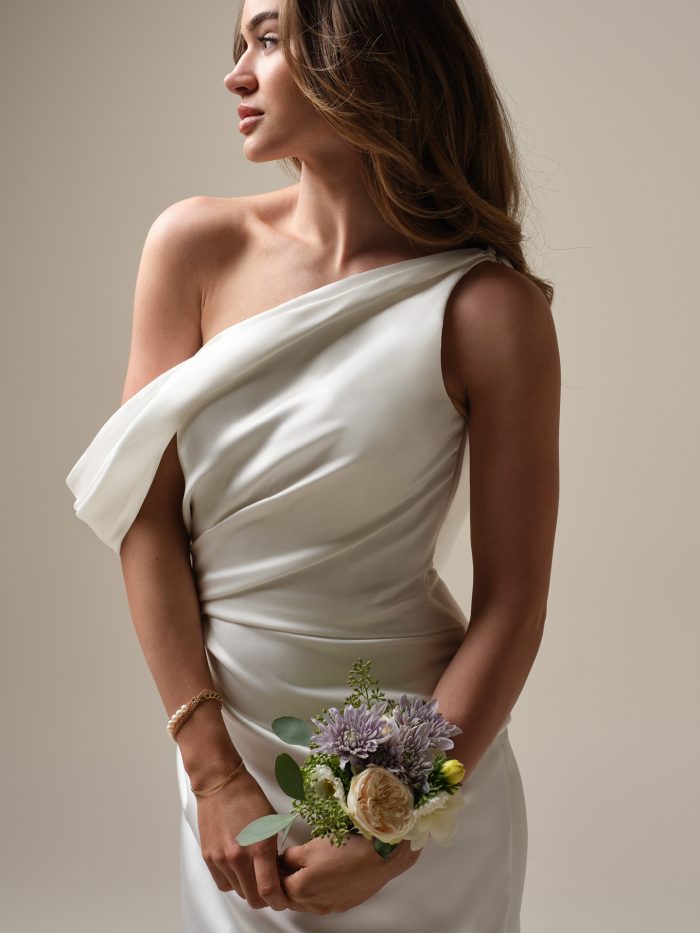 Close up of the Nadia gown showcasing the one shoulder draped satin neckline, soft ruching, and smooth fitted silhouette as the bride holds a small bouquet.