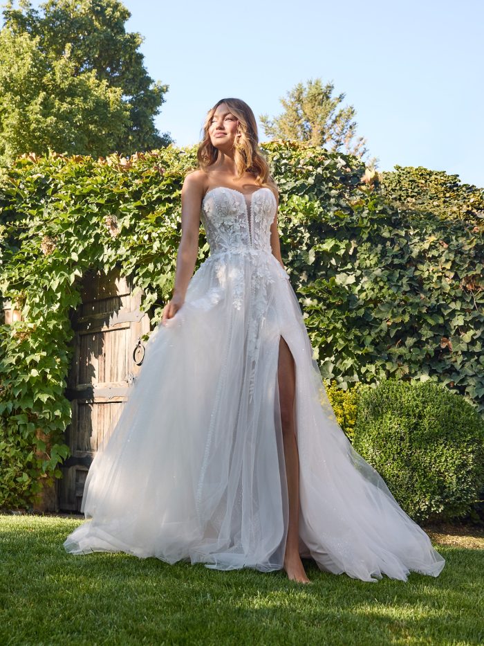 Bride wearing the Nora strapless A-line wedding dress featuring a sweetheart neckline, illusion corset bodice, beaded and sequin lace 3D motifs, flowing tulle skirt, and thigh-high slit, photographed outdoors in natural greenery.