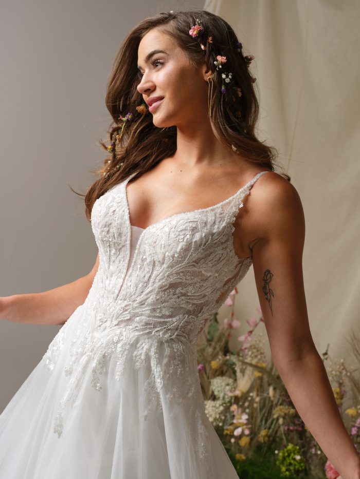 Close-up of the Valarie wedding dress by Rebecca Ingram, showcasing the intricate beaded lace bodice, illusion details, and delicate straps, with soft tulle skirt and floral hair accessories for a romantic bridal look.