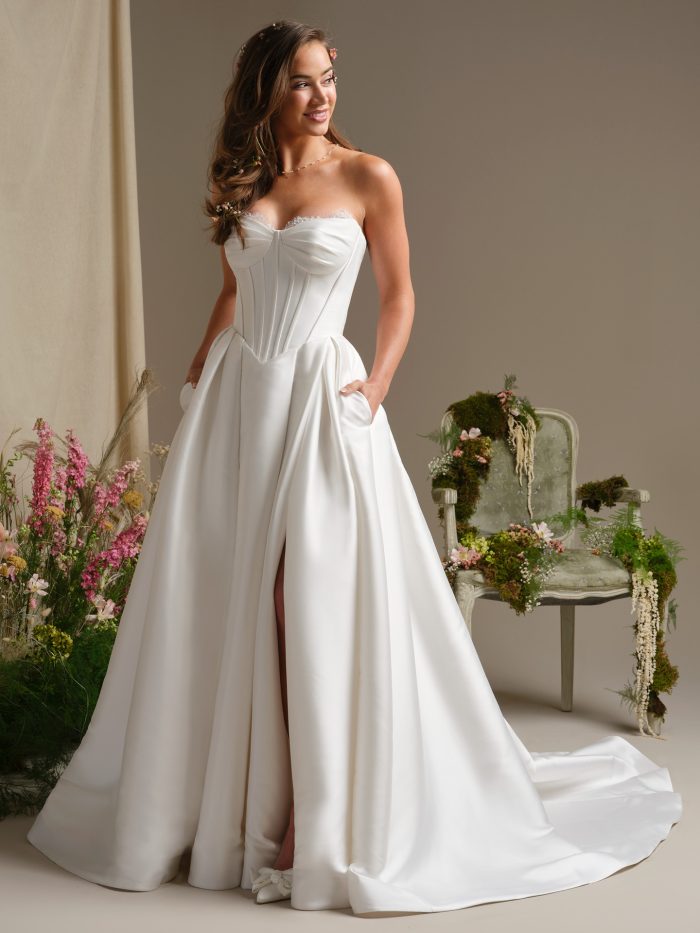 A model wearing the Rebecca Ingram Zorina wedding dress, featuring a structured strapless corset bodice with lace trim, a full satin ball gown skirt with a high front slit and hidden pockets.