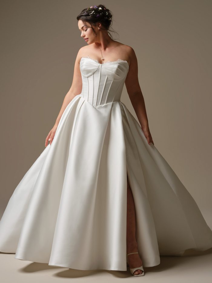 Curve model wearing Zorina by Rebecca Ingram — strapless satin ballgown wedding dress with exposed boning, soft pleated skirt, and thigh-high slit, styled with a romantic updo adorned with flowers.