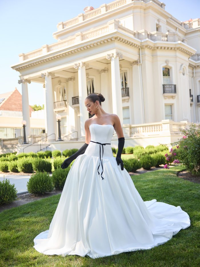 Bride wearing Adair by Sottero and Midgley, a strapless satin ball gown wedding dress with a clean neckline, full skirt, and black velvet ribbon belt, styled with long black gloves in front of a grand estate.
