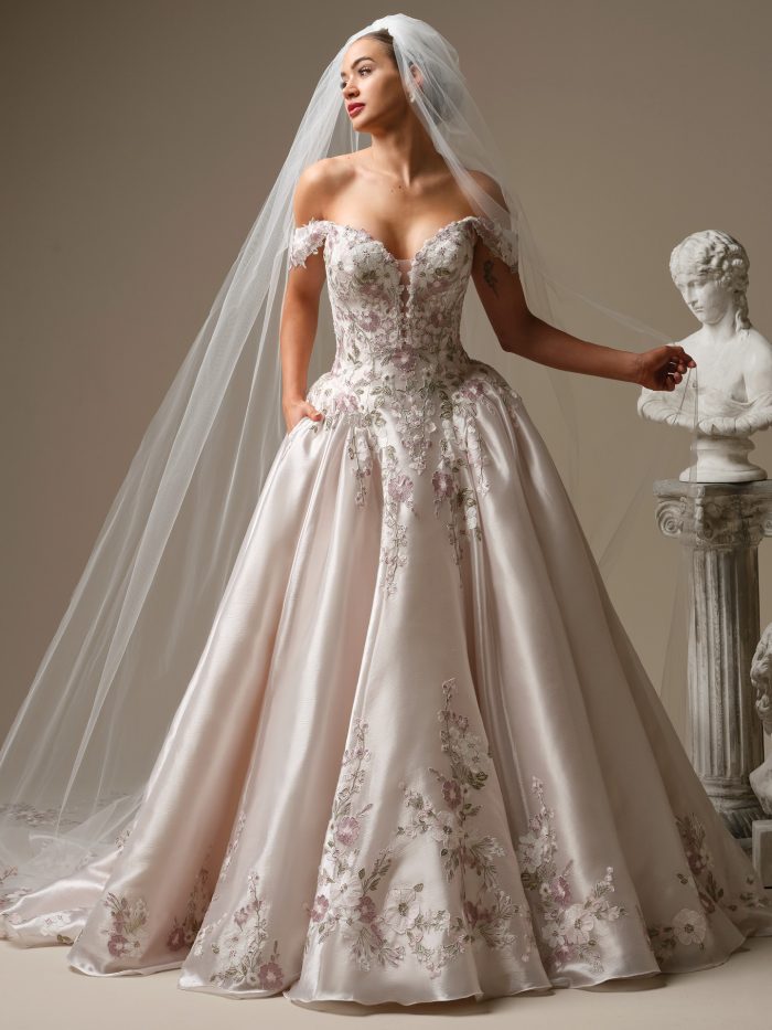 Bride wearing the Sottero and Midgley Alvarez ballgown wedding dress featuring an off-the-shoulder floral lace bodice with a plunging neckline, soft pink satin skirt with embroidered floral appliqués, and cathedral-length veil in a studio setting with classical bust statue.