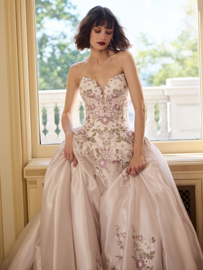 Close-up of a bride in a pink satin ballgown wedding dress featuring a strapless sweetheart neckline and embroidered floral appliqués. She poses by a window, highlighting the gown’s soft shimmer, romantic floral details, and structured corset bodice.