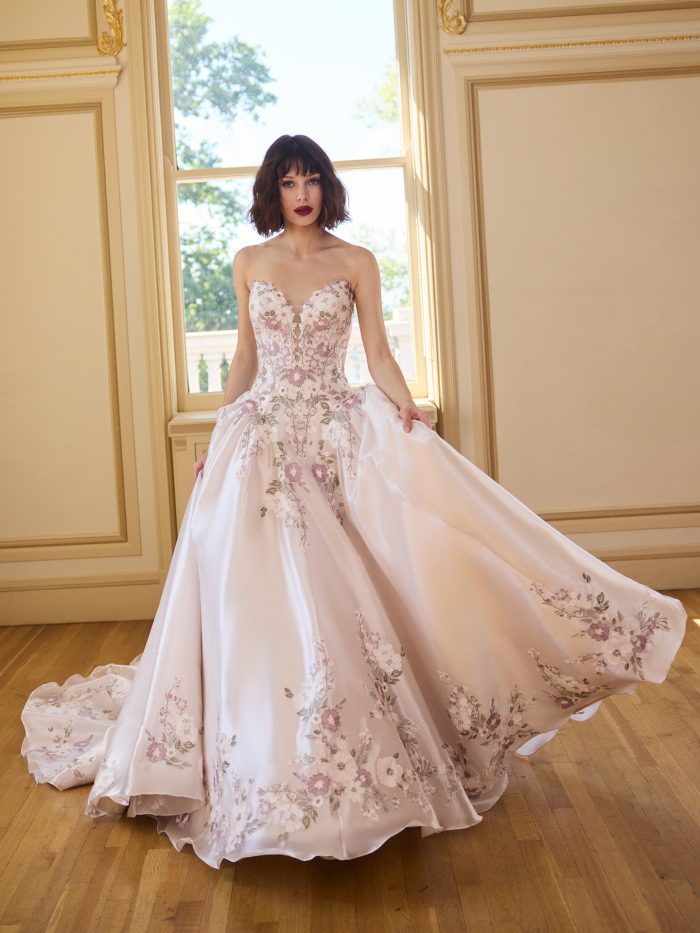 Bride wearing a pink satin ballgown wedding dress with embroidered floral lace and a strapless sweetheart neckline. The gown features dimensional lavender and ivory blooms with a full skirt and chapel train, creating a statement fairytale bridal look.