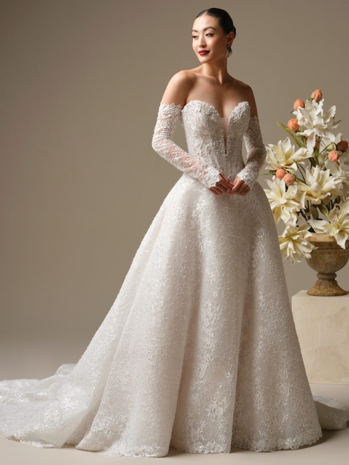 Angelica A-line wedding dress featuring off the shoulder lace sleeves, a deep illusion plunge, and a fully beaded floral lace skirt captured in a neutral studio setting.