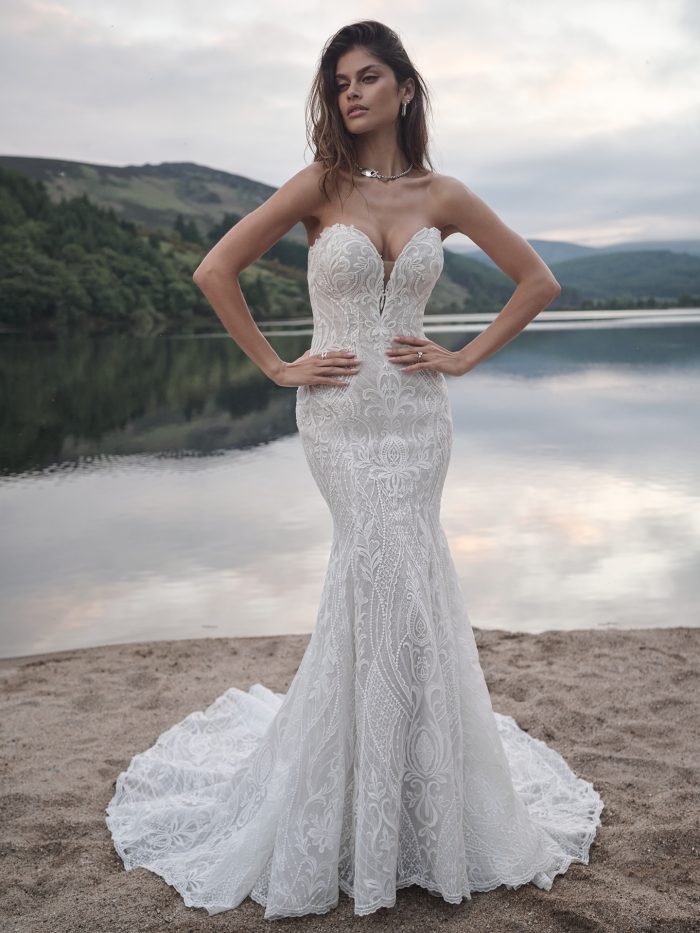Model wearing the Bronson sheath wedding dress, featuring bold allover lace, a plunging sweetheart neckline, and a fitted silhouette that flares into a dramatic lace train, photographed lakeside.