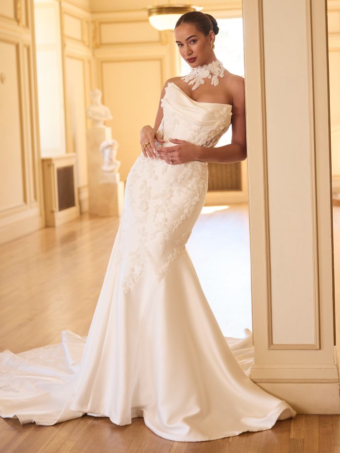Bride wearing the Sottero and Midgley Burke fit-and-flare wedding dress featuring a strapless draped bodice, curve-hugging silhouette, and detachable high-neck lace detail with floral appliqué.
