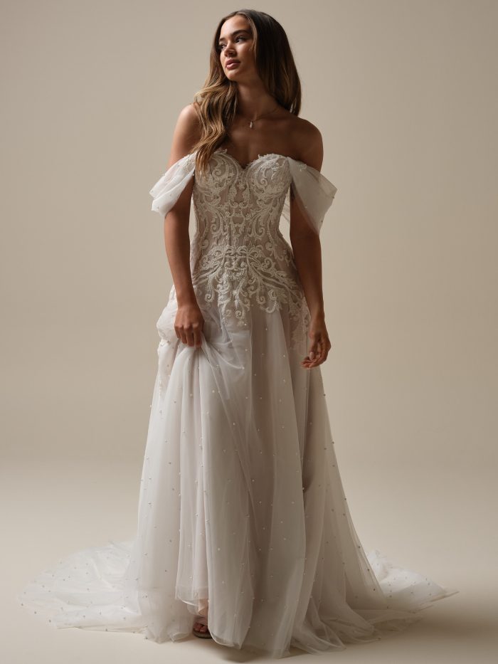 Bride wearing an off-the-shoulder beaded lace A-line wedding dress with illusion bodice and soft tulle skirt adorned with pearls, posing in soft studio lighting.