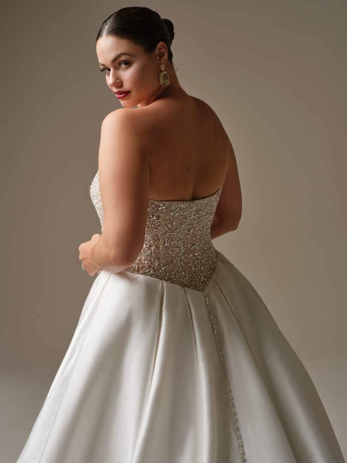Back view of Dmara by Sottero and Midgley featuring a strapless beaded bodice, crystal buttons trailing down the skirt, and dramatic satin ball gown silhouette.