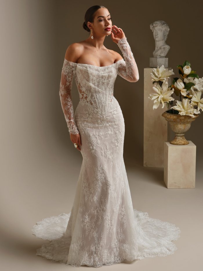 Bride wearing a fit-and-flare lace wedding dress featuring an off-the-shoulder neckline, exposed boning, and illusion lace sleeves. The gown is adorned with delicate beading and floral appliqués, styled with bold earrings for a modern, romantic bridal look.