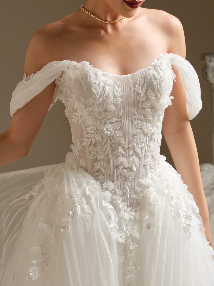 Close-up of a bride wearing an off-the-shoulder ballgown wedding dress with pleated tulle sleeves and 3D floral lace appliqués. The structured bodice features exposed boning and shimmering beadwork for a romantic, couture-inspired bridal look.