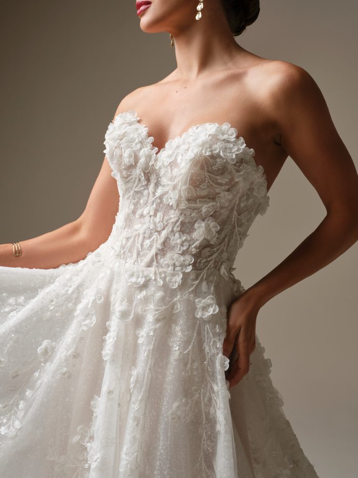 Close-up of a strapless sweetheart ballgown wedding dress with dimensional floral lace and sequined tulle. The bride gently lifts the gown’s skirt, highlighting the sheer corset bodice and intricate floral detailing.