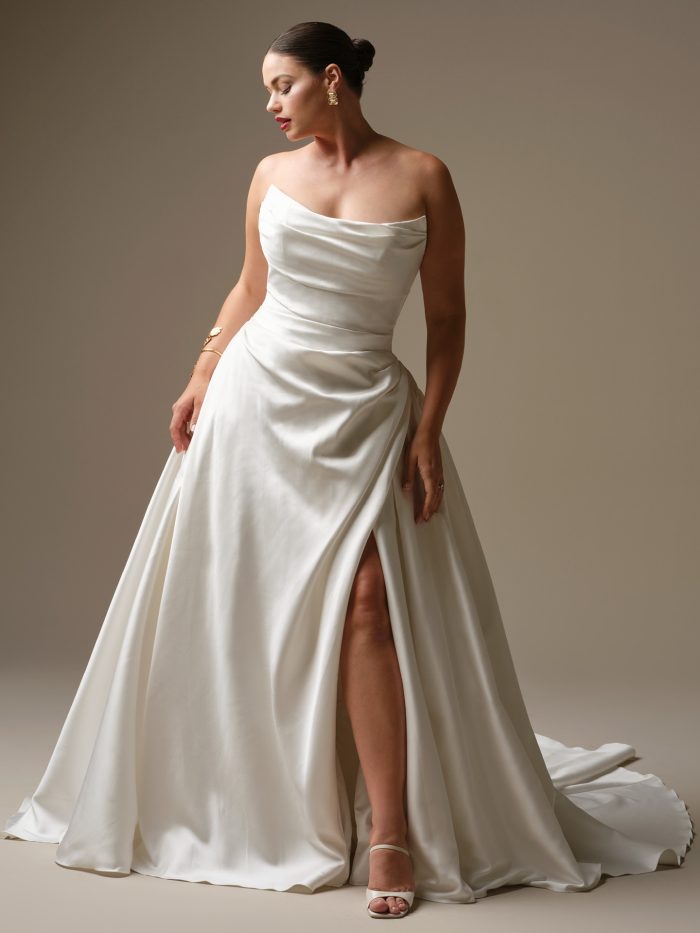 Curve model wearing Kincaid by Sottero and Midgley — modern satin ballgown wedding dress with asymmetrical neckline, ruched bodice, and thigh-high slit, styled with gold accessories and sleek bun.