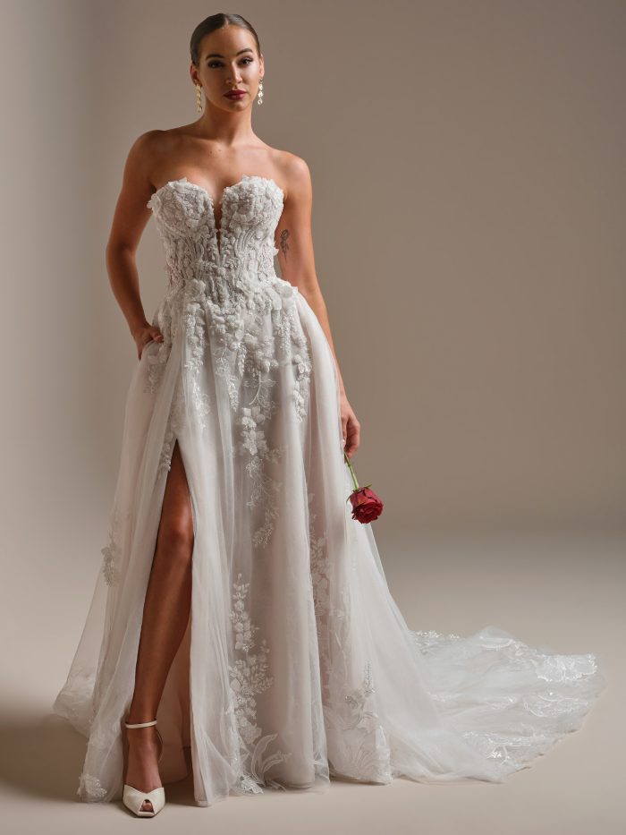 Bride wearing a strapless A-line wedding dress with a plunging neckline, 3D floral lace bodice, and high slit skirt. The gown features sheer tulle layers and beaded embroidery, styled with statement earrings and a single red rose for a modern romantic look.