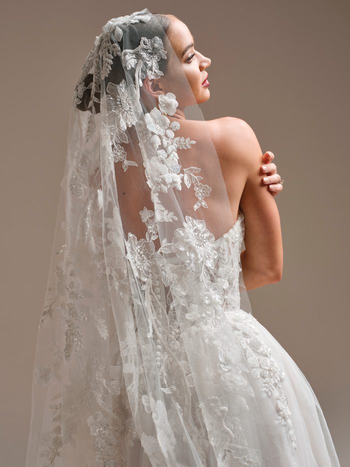 Bride wearing a cathedral-length veil adorned with dimensional floral lace appliqués and shimmering embroidery. The veil drapes over a lace wedding dress, creating a soft, romantic look with intricate texture and elegant floral detailing.