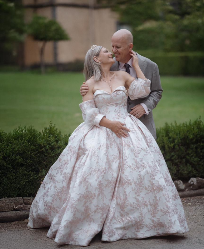 Bride in a patterned off-the-shoulder ball gown with detachable sleeves and tiara, smiling and embracing groom in a gray suit in an outdoor garden setting.