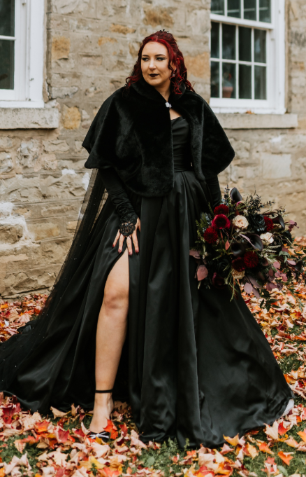 Bride in a dramatic black wedding dress with a slit skirt, lace sleeves, and velvet cape, holding a dark floral bouquet while standing on autumn leaves outside a rustic stone building.