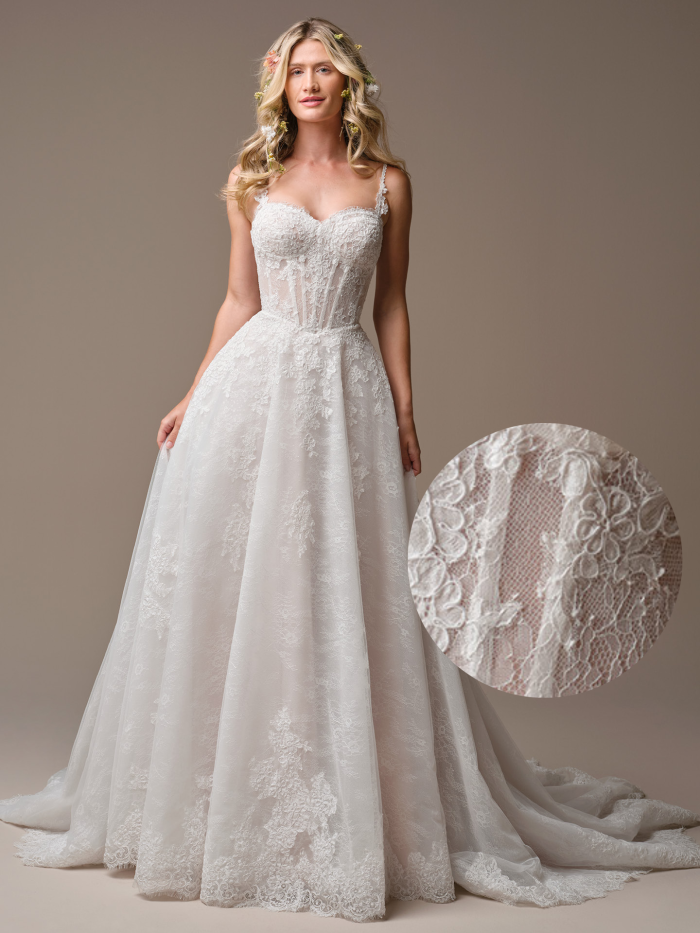 Bride wearing the Esmeralda by Rebecca Ingram, an A-line lace wedding dress with a corset-style bodice, sweetheart neckline, delicate lace straps, and a full flowing skirt with floral lace appliqués, styled with loose waves and floral hair accents in a neutral studio setting.